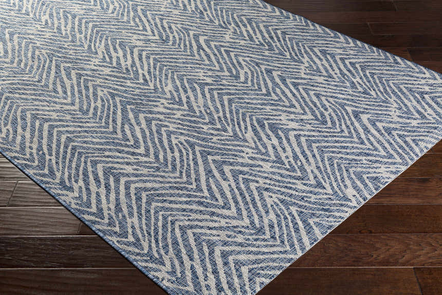Close-up of the Ashcamp Area Rug. Blue and taupe chevron pattern on a rectangular rug, on dark wood flooring. Corner