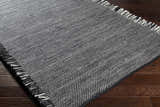 Close-up of the hand-woven Boyden Black Area Rug with Tassels. Black and cream recycled PET yarn on dark wood floor. Corner