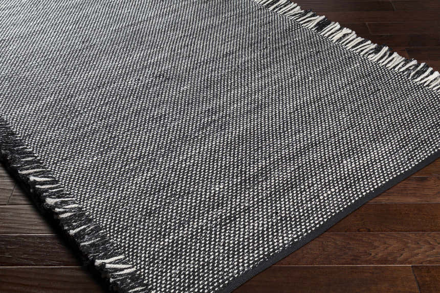 Close-up of the hand-woven Boyden Black Area Rug with Tassels. Black and cream recycled PET yarn on dark wood floor. Corner