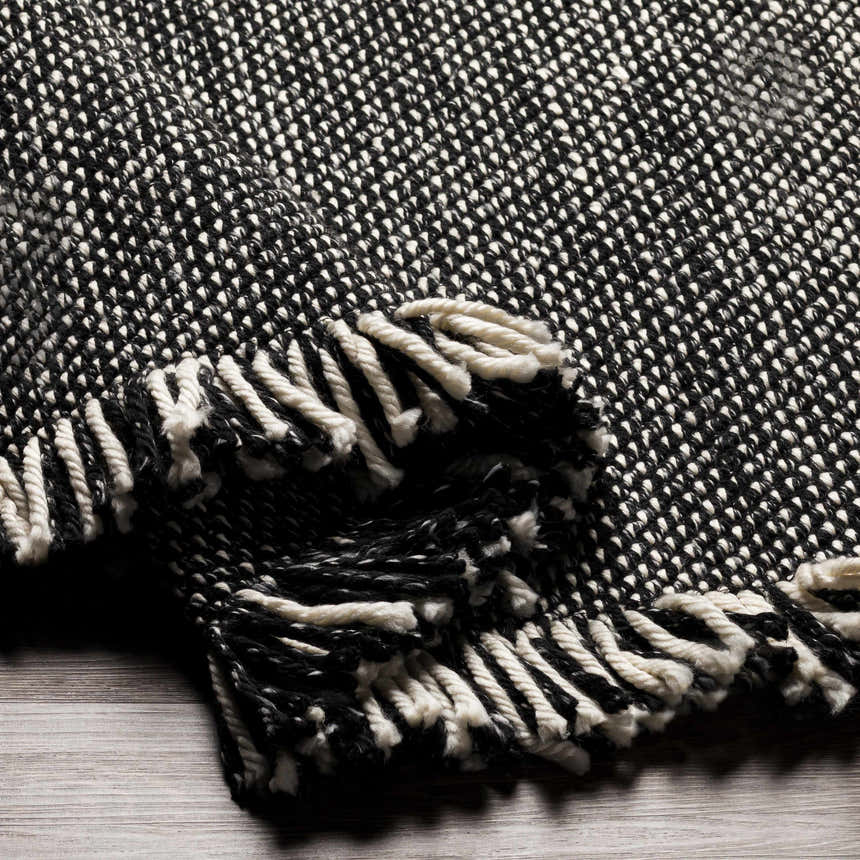 Close-up of the Boyden Black Area Rug with Tassels. Hand-woven texture in black and cream recycled yarn, with knotted fringe. Fold