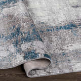 Close-up of the Bugho Area Rug showing its cream, gray, and teal abstract design and soft, textured polyester material. Fold
