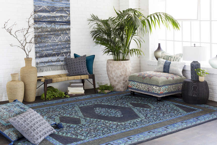 Living room scene with a Chesham Clearance Rug in charcoal, blue, and teal tones, styled with a bench, ottoman, and potted plant. Roomscene-Rectangle