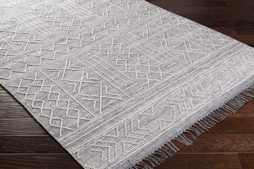 Close up of Gray Clintwood Wool Tassel Carpet on dark wood floor. Bohemian style rug with geometric pattern. Corner