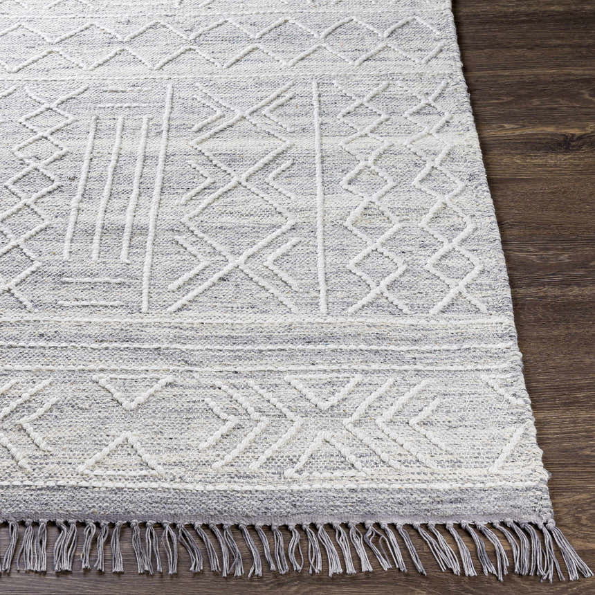 Close-up of Gray Clintwood Wool Tassel Carpet on dark wood floor. Charcoal, cream, and white geometric pattern. Hand woven texture. Front