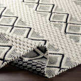 Close-up of the hand-woven Cobourg Area Carpet. Black geometric diamonds on a cream wool rug with a textured, looped pile. Fold
