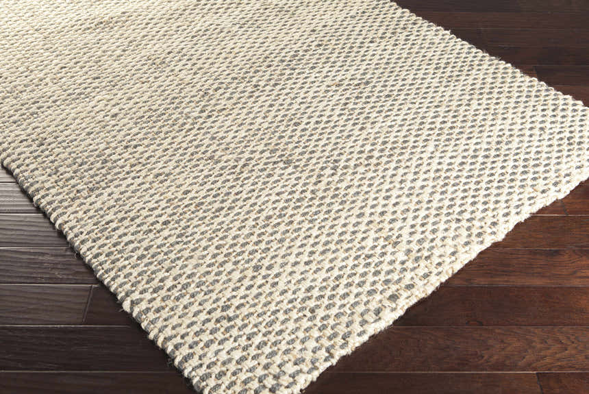 Close-up of a hand-woven Carrier Clearance Jute Carpet. Natural fiber rug with charcoal and ivory woven pattern on dark wood floor. Corner