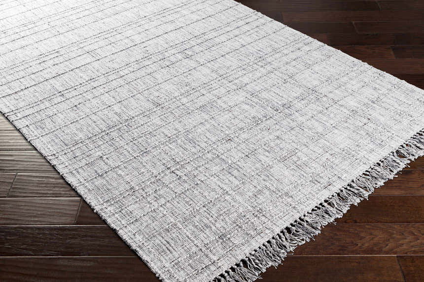 Close-up of the hand-loomed Delanco Area Rug, showing its ivory and dark brown weave and fringe on dark brown wood floors. Corner