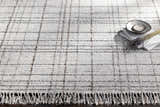 Close up of the hand-loomed Delanco Area Rug. Features a contemporary dark brown and pale blue plaid design with white fringe. Styleshot