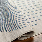 Close-up of the Duri Area Rug. Features a geometric pattern with admiral blue lines on a natural gray background. Fold