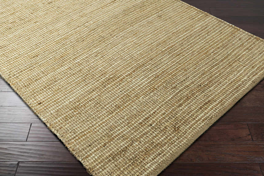 Close-up of the Frederic Jute Carpet showing its tan color, hand woven texture, and rectangular shape on dark wood flooring. Corner