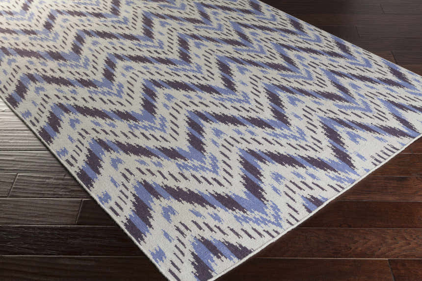 Close-up of Fryeburg Area Rug: hand-woven wool rug with beige ground and lavender chevron pattern on dark wood floor. Corner