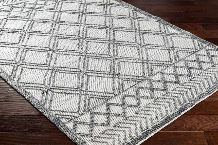 Close-up of the Gada Area Rug, a hand-woven rug with a geometric dark gray and light gray diamond pattern on wood floors. Corner