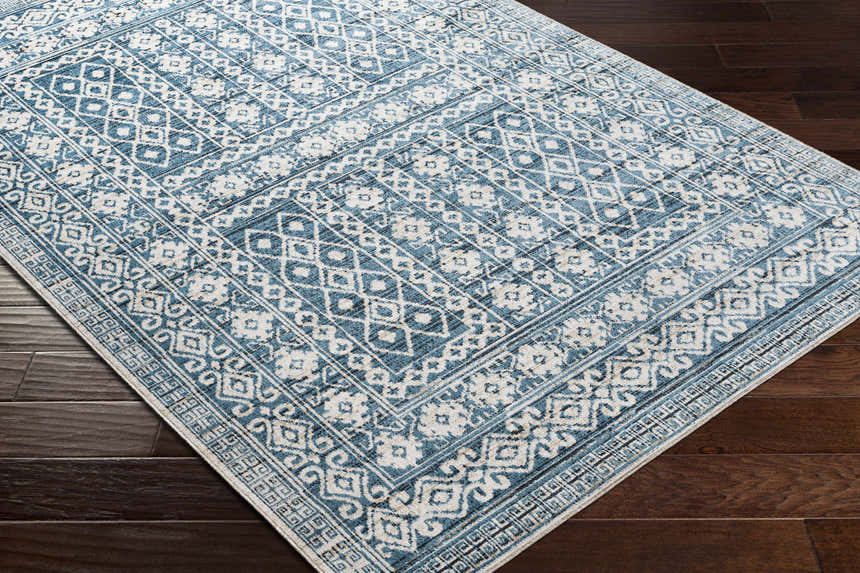 Close-up of the Gandy Washable Area Rug. Features a blue and cream geometric pattern on a dark wood floor. Corner