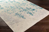 Close-up of Sweeny Beige/Blue Abstract Area Rug on dark wood floor. Beige rug with teal and slate abstract pattern. Corner