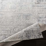 Close-up of Hardeeville Area Rug: a textured, abstract pattern in shades of gray and white on dark wood. Fold