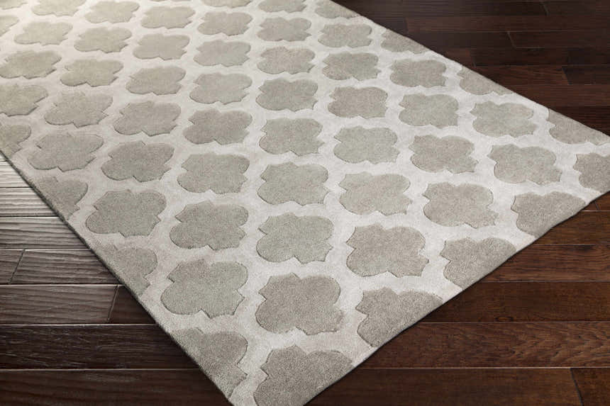 Close-up of the Highspire Area Rug. Light gray rug with a taupe quatrefoil pattern on dark wood flooring. Corner