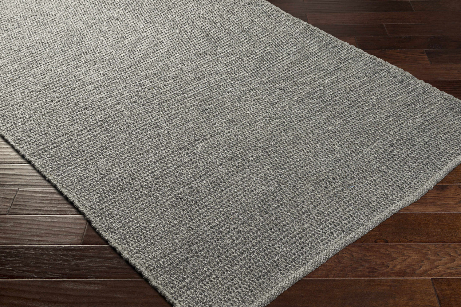 Close-up of the Innisfail Area Rug, a hand-woven rug in medium gray, showcasing its texture and construction on wood floors. Corner
