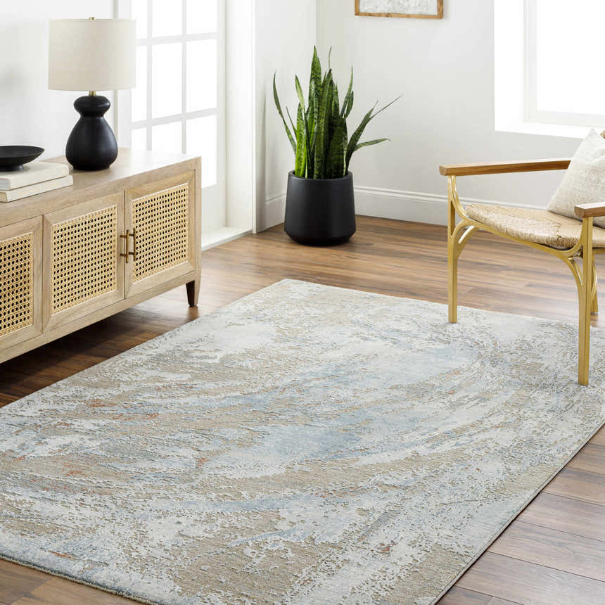 Living room scene with a Jaela Thick Luxe Area Rug in gray, brown, and blue tones. The rug is styled with a wood cabinet and woven chair. Roomscene-Rectangle