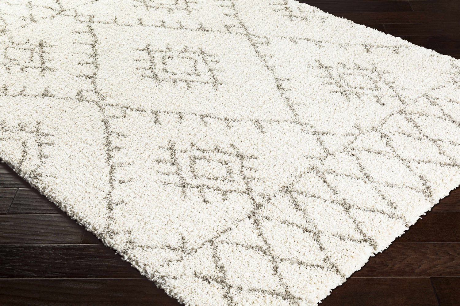 Close-up of the ivory Kunia Area Rug, featuring a geometric shag design on dark wood flooring. Corner