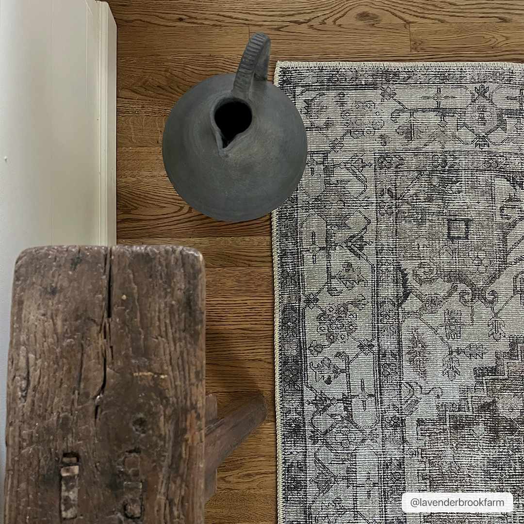 Close-up of Athor Washable Rug in dark gray, eggplant, and natural gray on wood floor with rustic wood stool and gray ceramic vase. Styleshot
