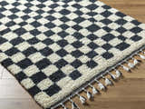 Close-up of the Uttam Area Rug, a hand-woven wool rug in a black and light silver checkerboard pattern with tassels. Corner