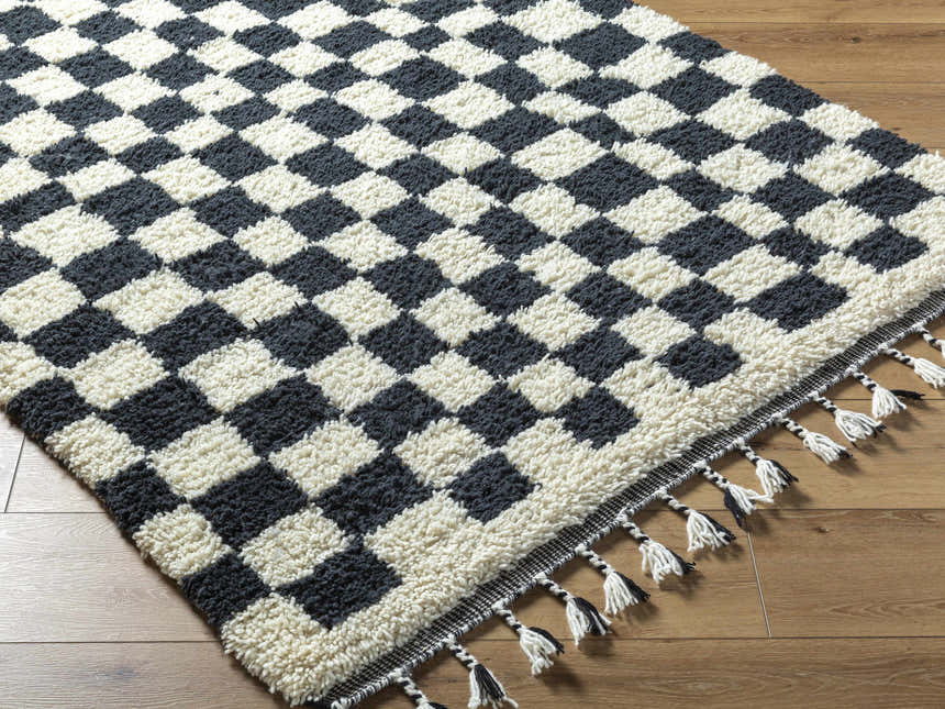 Close-up of the Uttam Area Rug, a hand-woven wool rug in a black and light silver checkerboard pattern with tassels. Corner
