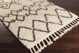 Close-up of the Lothian Area Rug. Ivory shag rug with charcoal geometric pattern and tassels on dark wood floor. Corner