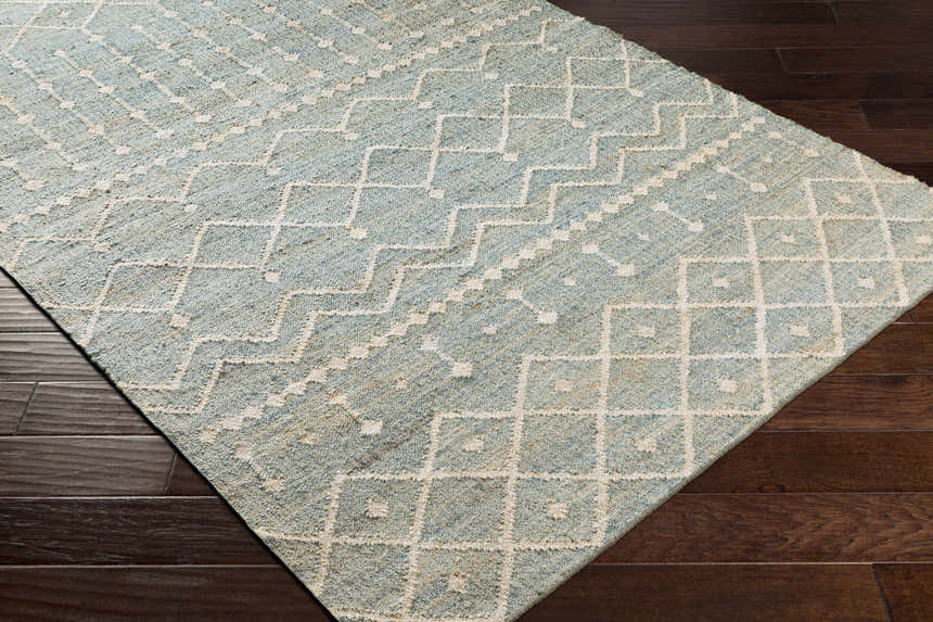 Close-up of the Lowville Jute Rug. Hand-woven rug in dusty sage with a geometric ivory pattern on a dark wood floor. Corner