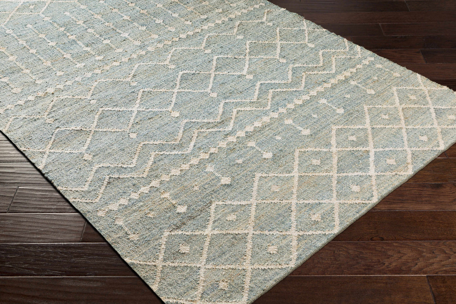Close-up of the Lowville Jute Rug. Hand-woven rug in dusty sage with a geometric ivory pattern on a dark wood floor. Corner