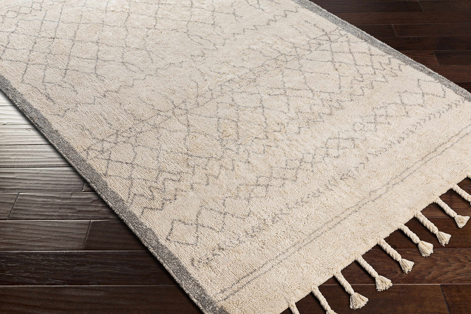 Close-up of the cream and gray Luncarty Area Rug with geometric pattern and tassels on dark wood floors. Corner