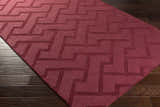 Close-up of the plum Magness Area Rug showing its geometric pattern and hand-loomed texture on a dark wood floor. Corner