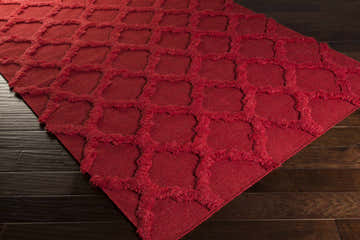 Close-up of the red Marilla Area Rug, a hand-woven shag rug with a geometric pattern, on a dark wood floor. Corner