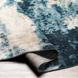 Close-up of the Masiga Washable Area Rug. Abstract design in shades of blue, beige, and black. Machine woven polyester rug. Fold