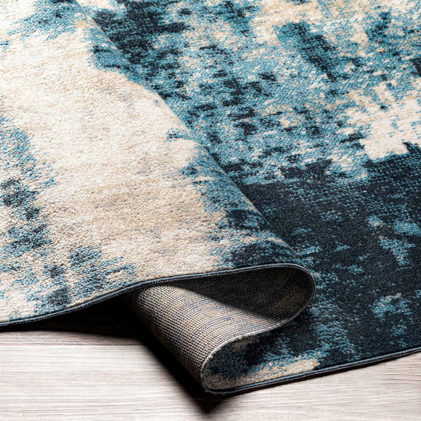 Close-up of the Masiga Washable Area Rug. Abstract design in shades of blue, beige, and black. Machine woven polyester rug. Fold