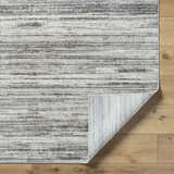 Close-up of the Munya Area Rug showing charcoal, cream, and gray horizontal stripes on a wood floor. Rug corner is folded. Fold
