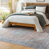 Bedroom scene with a beige and blue Mianay Washable Area Rug, wood bed, gray bedding, and natural light. Roomscene-Rectangle