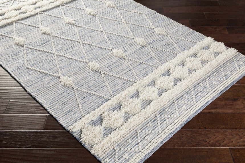 Close up of the Moe Area Rug, a hand woven rug with a blue and cream diamond pattern and textured details. Corner