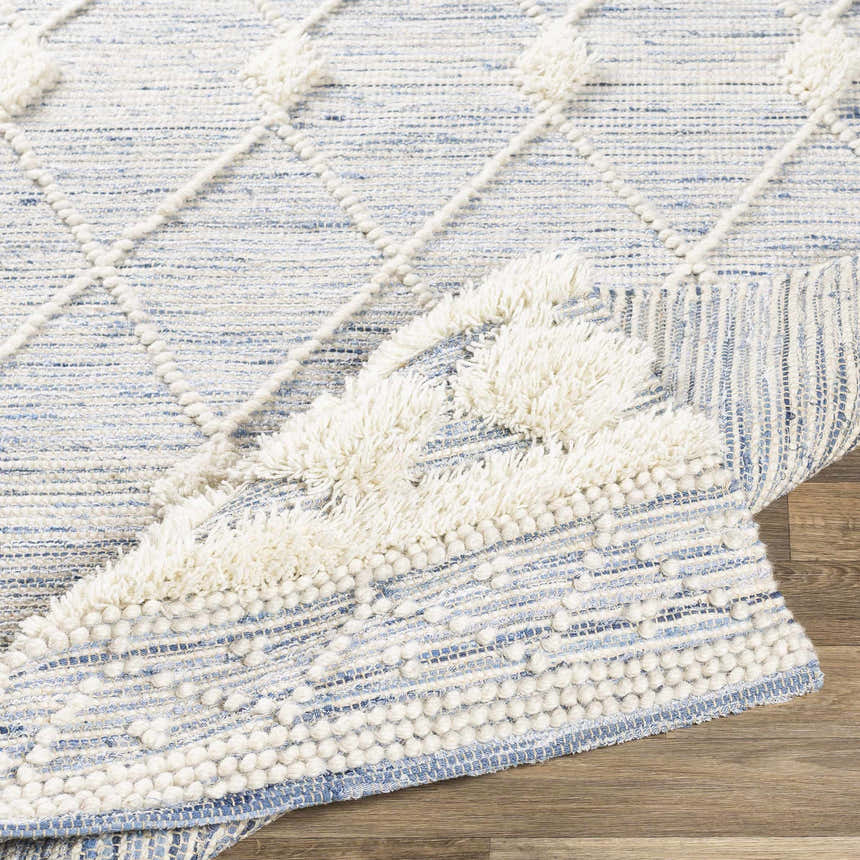 Close-up of the Moe Area Rug. Hand-woven rug in blue and cream with a diamond pattern and fringe detail. Fold