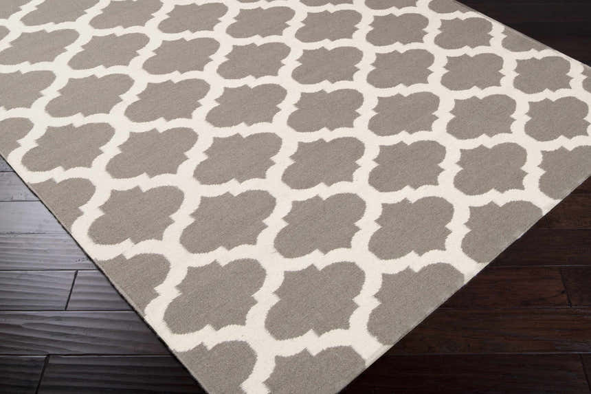 Close-up of the Neshkoro Wool Area Rug. Gray and light beige quatrefoil pattern on a hand-woven rug with dark wood floors. Corner