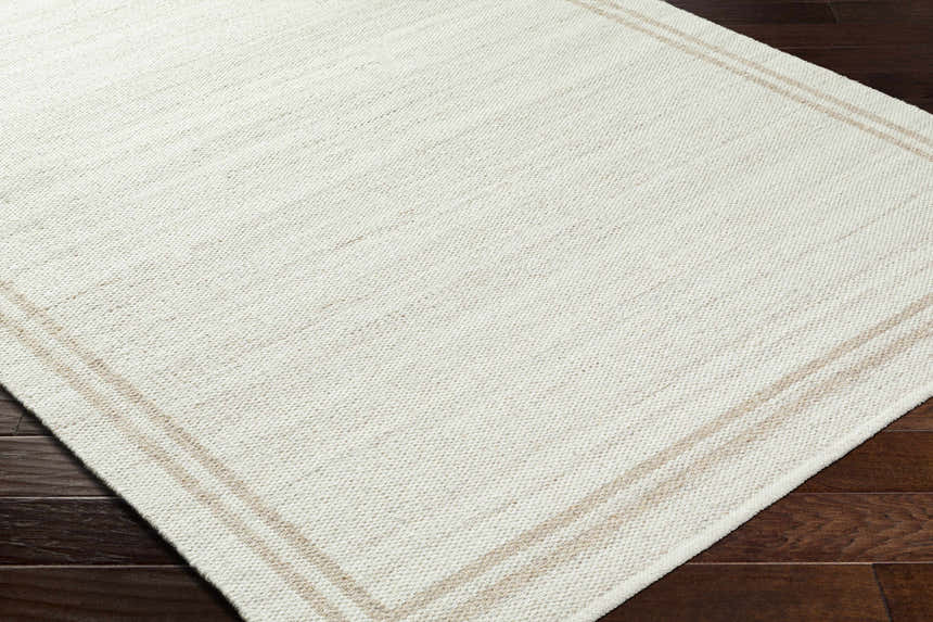 Close-up of the hand-woven Nimue Area Rug. Ivory wool rug with a simple tan border on dark wood flooring. Corner