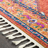 Close-up of the Oban Traditional Rug showing camel, coral, and blue hues, plus a detailed pattern and braided fringe. Texture