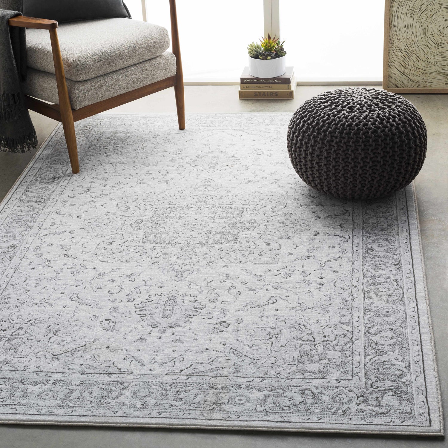 Living room scene with a light gray Okehampton Area Rug, charcoal pouf, and modern chair. Roomscene-Rectangle