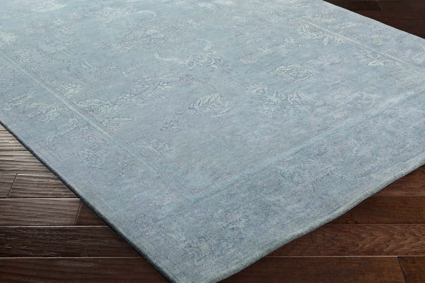 Close-up of the Oxly Area Rug showing a pale blue wool rug with a traditional floral pattern on dark wood floors. Corner