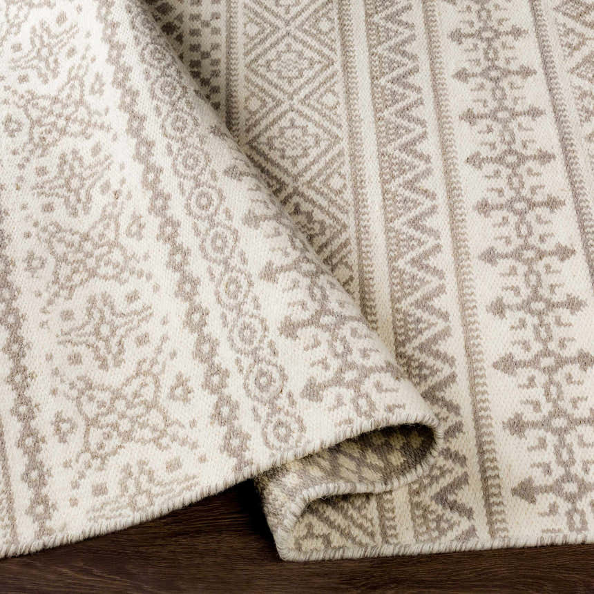 Close-up of the hand-woven Pumwani Area Carpet. Cream wool rug with gray bohemian-style pattern and fringe detail. Fold