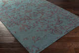 Close-up of the hand-knotted Redvale Area Rug. Teal rug with a burgundy, contemporary pattern on a dark wood floor. Corner
