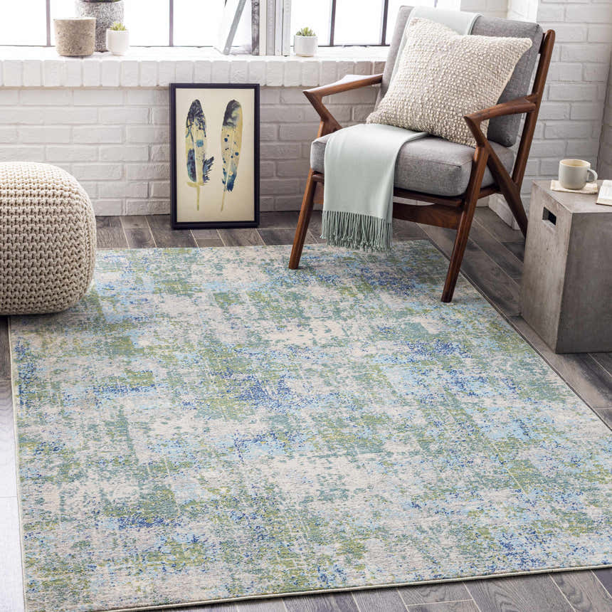 Coastal living room scene with a Riegelwood Area Rug in light beige, green, and blue tones. Gray chair and woven pouf. Roomscene-Rectangle