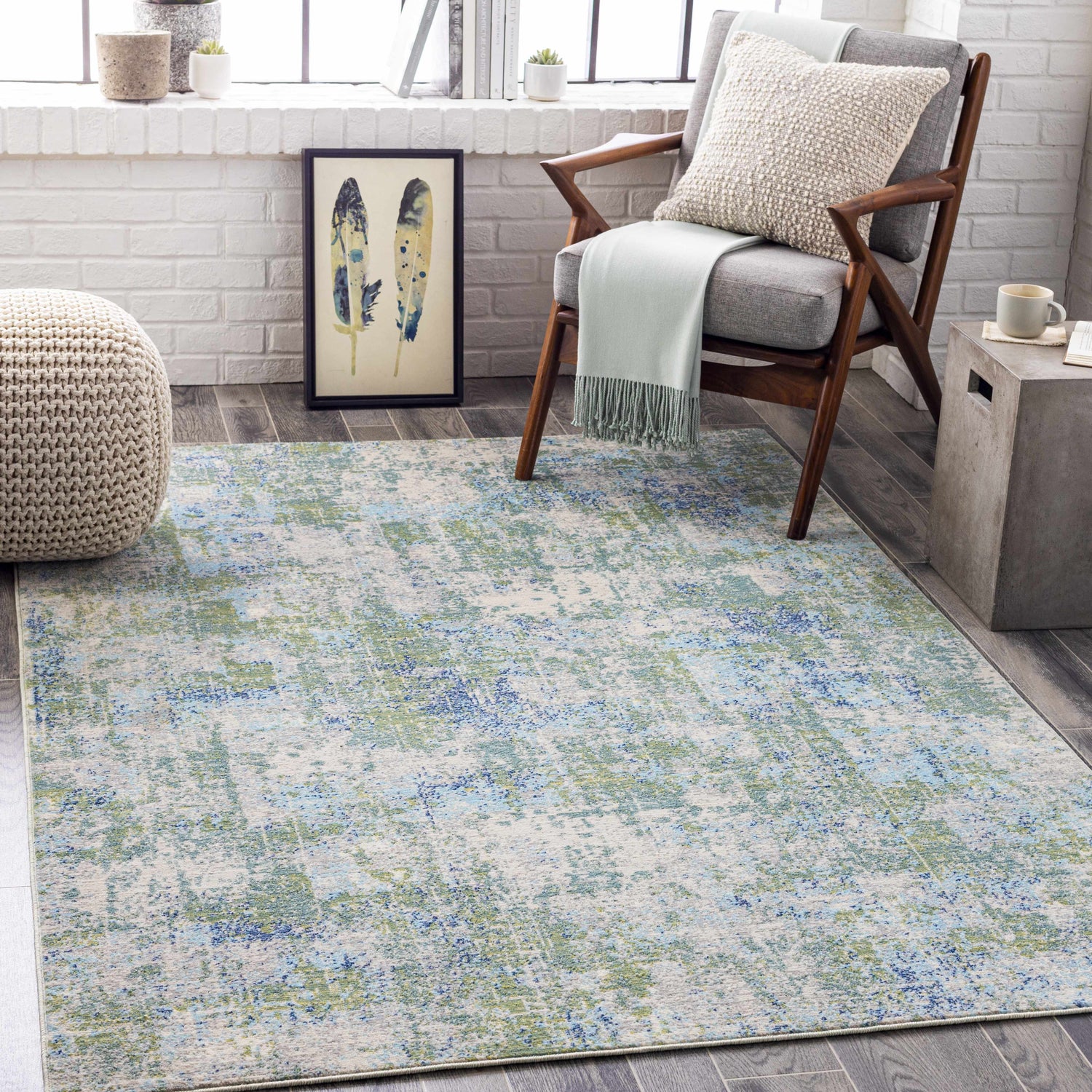 Coastal living room scene with a Riegelwood Area Rug in light beige, green, and blue tones. Gray chair and woven pouf. Roomscene-Rectangle