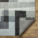 Close-up of Raisa Area Rug showing geometric pattern in aqua, black, cream, and gray on wood floor. Machine woven texture detail. Fold