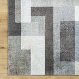 Close-up of the Raisa Area Rug showing a contemporary geometric pattern in cream, gray, and black on a wood floor. Corner