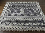 Hand woven Rossville Area Rug on dark wood floor. Features a black and white geometric design with fringe. Front